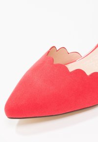 Chaussure plate en daim rouge à bout pointu avec détail de bord festonné et petit talon, présentée sur fond blanc.