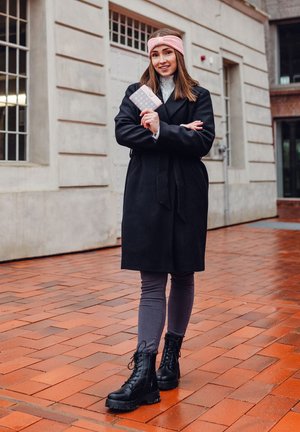 Black coat with belt, gray leggings, and black lace-up boots; holding a light pink wallet. Background features brick pavement and building.