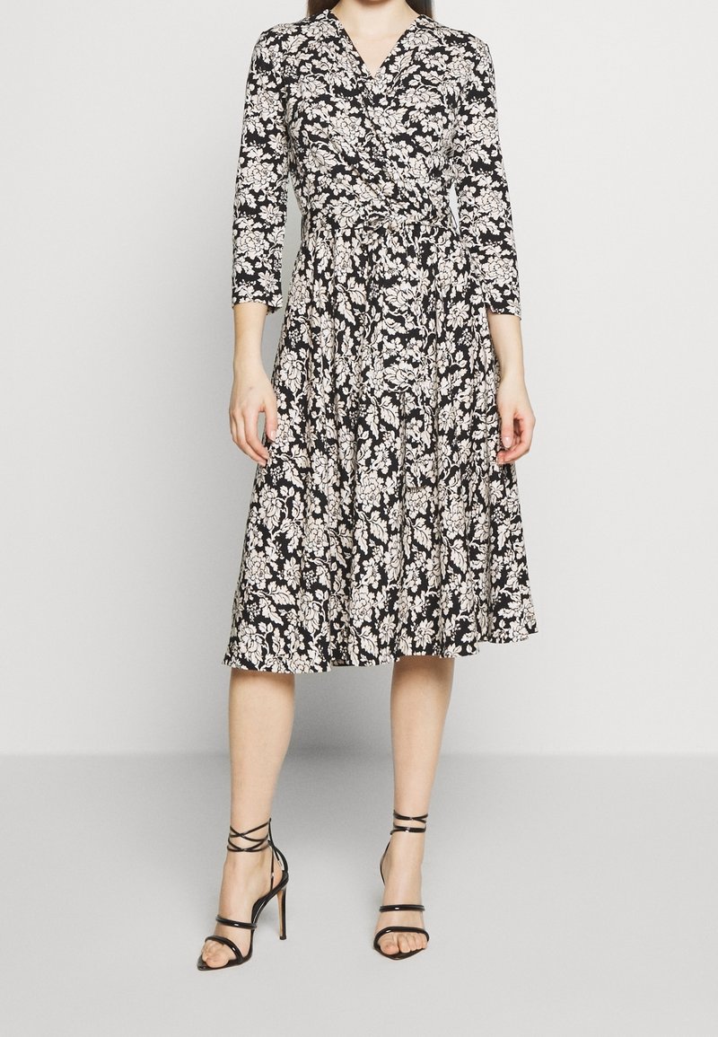 Femme portant une robe portefeuille noire à imprimé floral blanc, manches trois-quarts, et des sandales noires à talons hauts à brides, debout devant un fond uni.