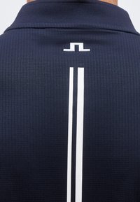 Πίσω μέρος πουκάμισου ναυτικού μπλε με υφή, με λευκό γεωμετρικό λογότυπο πάνω από δύο κάθετες λευκές ρίγες που τρέχουν κατά μήκος του κέντρου.