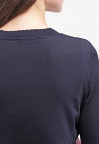 Top a maniche lunghe blu navy a costine con scollo rotondo e design aderente, che mostra un tessuto testurizzato dalla vista laterale.