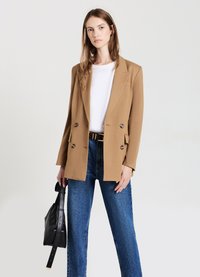 Blazer doppiopetto color cammello con rever a crestina, due tasche frontali e bottoni dorati, abbinato a jeans a vita alta e una borsa nera.