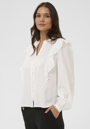 Žena s dlhými hnedými vlasmi, oblečená v bielej blúzke s volánikmi a gombíkovými detailmi a čiernych nohaviciach, stojaca s jednou rukou v vrecku.