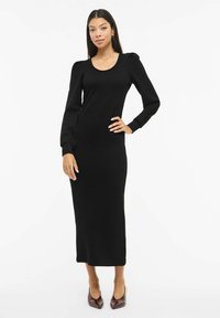 Musta pitkähihainen neulemekko, jossa on pyöreä pääntie, istuva siluetti ja muhkeat olkapäädetaljit, yhdistettynä ruskeisiin, teräväkärkisiin korkokenkiin.