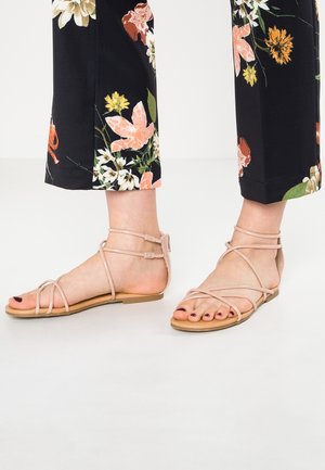 Feet wearing nude strappy flat sandals paired with black pants featuring orange, white, and green floral patterns.
