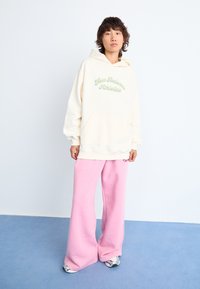 Sweatshirt à capuche crème avec "New Balance Athletics" en texte vert, associé à un pantalon de jogging léger et ample rose pâle. Chaussures de sport grises visibles.