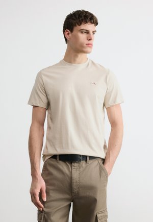 Mlad moški, oblečen v bež majico s kratkimi rokavi z majhnim rdečim logotipom CK in kaki hlače z žepi, stoji z eno roko v žepu.