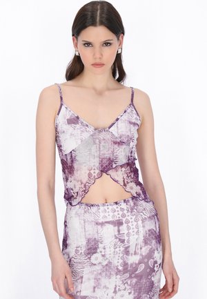 Tânără cu păr castaniu drept poartă un top crop fără mâneci mov cu alb, cu model, și o fustă asortată, stând pe un fundal alb.