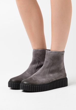 Nohy ve šedých semišových kotníkových botách s tlustými černými podrážkami na bílém pozadí.