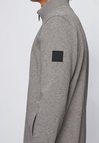 Grå zip-up sweatshirt med hög krage, sidoslitsar och en svart fyrkantig logotyp på vänster axel. Mjuk tygkvalitet.