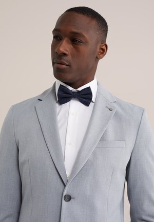 Man wearing light gray suit jacket, white dress shirt, and dark blue polka dot bow tie, looking to the side against plain background.