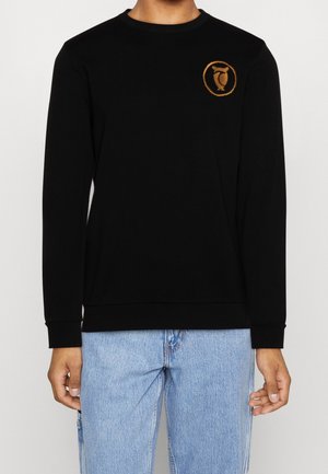 Persona che indossa una maglietta nera a maniche lunghe con un piccolo logo arancione circolare a forma di gufo sul petto sinistro e jeans azzurri.
