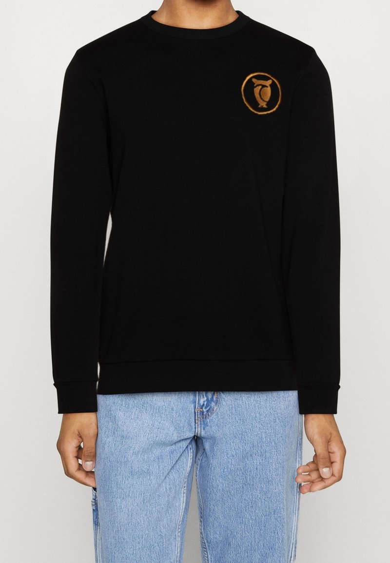 Personne portant un t-shirt noir à manches longues avec un petit logo circulaire orange de hibou sur la poitrine gauche et un jean bleu clair.
