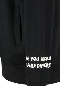 Schwarzer Pullover mit geripptem Saum und Bündchen. Auf dem Ärmel befindet sich ein weißer Aufdruck mit dem Text: „ICH MAG DICH, WEIL DU ANDERS BIST.“