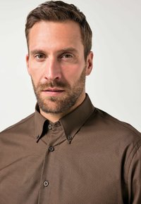 Hombre barbudo con cabello marrón corto que lleva una camisa de botones marrón, mirando directamente a la cámara con una expresión neutral.