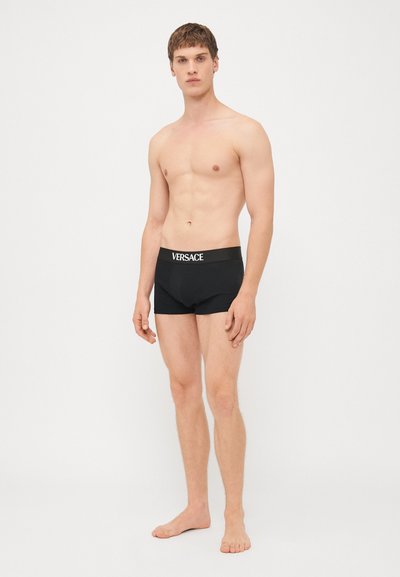Schwarze Boxershorts aus einem glatten Stoff mit einem elastischen Taillenbund, der "VERSACE" in weißen Buchstaben zeigt. Das Modell posiert barfuß.