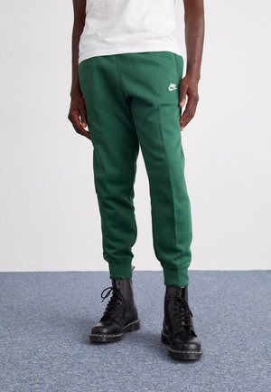 Grüne Baumwoll-Jogginghosen mit elastischen Bündchen, die mit einem kleinen weißen Nike-Logo am Oberschenkel versehen sind, kombiniert mit schwarzen Schnürstiefeln.