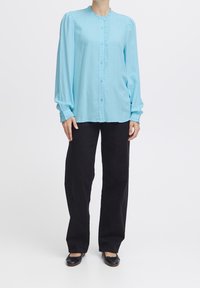 Camicia azzurra con bottoni e maniche lunghe, con parte anteriore arricciata, scollo rotondo, e pantaloni neri a vita alta. Indossata con scarpe flat nere.