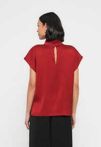 Punainen satiinitoppi, jossa on korkea kaulus, liehukkeet olkapäillä ja avonainen takakappale. Sileä pinta ja väljä, rento istuvuus.