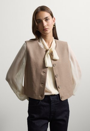 Giovane donna con lunghi capelli castani indossa un gilet beige con bottoni sopra una camicetta bianca con fiocco davanti e pantaloni neri, posando davanti a uno sfondo semplice.
