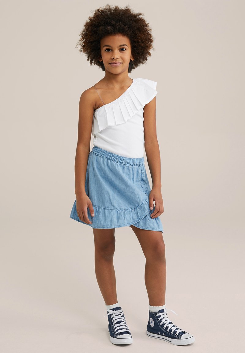 Weißes Tops mit einem Schulterträger und Rüsche, kombiniert mit einem hellen Jeansrock mit ausgestelltem Saum. Marineblaue High-Top-Sneaker runden das Outfit ab.