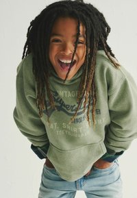 Sudadera verde con un estampado gráfico, vaqueros de mezclilla y dreadlocks largos y texturizados; el sujeto sonríe ampliamente contra un fondo liso.