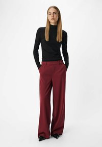 Zwarte aangesloten coltrui gecombineerd met burgundy wijde broek. De broek heeft een voorplooi; het model draagt puntige zwarte schoenen.