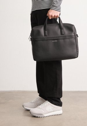 Personne portant un pantalon noir et des baskets blanches tenant une mallette en cuir noire Calvin Klein avec deux poignées.