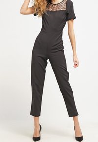 Svart jumpsuit med korta ärmar, spetsdetalj vid halsringningen, smalnande ben och en mjuk textur. Tillsammans med svarta högklackade skor.