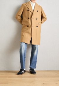 Cappotto doppiopetto beige in misto lana con bottoni neri, abbinato a jeans a gamba dritta blu sbiaditi e sneakers nere con strisce bianche.