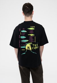 Black oversized cotton t-shirt featuring colorful text and graphics of a figure and tour dates from "The Cure's" 1986 Beach Party Tour.