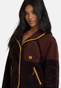 Polarowa kurtka w kolorze bordowym z czarnymi wzorami, z żółtymi akcentami, przednimi kieszeniami na zamek oraz małą kieszonką na piersi. Teksturowany materiał.