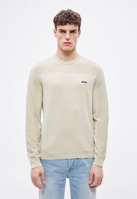 Bézs pulóver bordázott textúrával az ujjakon és a felső mellrészen. A bal oldalon egy kis fekete "BOSS" logó található. Klasszikus kerek nyakkivágás.