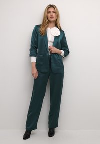 Teal satīna žakete ar divām priekšējām kabatām, uzvilkta pār baltu krekli, kas izdaiļots ar teksturētu ziedu akcentu, apvienota ar saskaņojošām biksēm un melniem potīšu zābakiem.