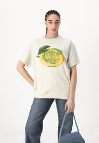 Camiseta oversized color crema con un gráfico de limón amarillo y el texto "BITTER SWEET" en verde. Combinada con unos vaqueros de mezclilla azul.