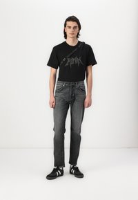 Zwarte grafische T-shirt met vetgedrukte tekst, gecombineerd met donkergrijze jeans en zwarte sneakers met witte strepen. Eenvoudig, casual ontwerp.