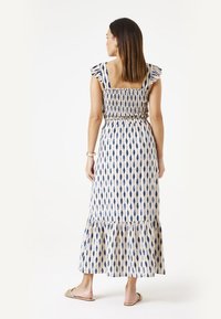 Maxi jurk in crème met blauw diamanten patroon. Heeft rimpelbandjes, een gerimpelde bovenkant en een gelaagde rok. Draagt bij platte sandalen.