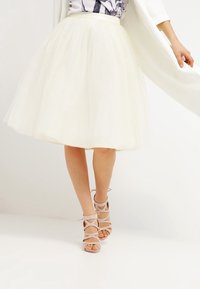 White tulle midi skirt with a defined waist, layered texture, worn with light pink strappy sandals featuring cut-out details.