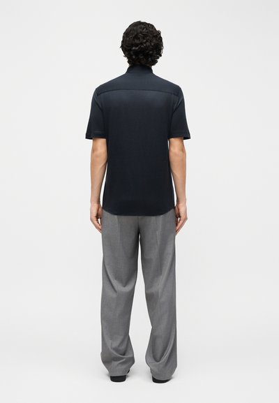 Άνδρας που στέκεται με την πλάτη στην κάμερα, φορώντας μαύρο κοντομάνικο πόλο και γκρι παντελόνια με φαρδύ πόδι, μπροστά σε απλό λευκό φόντο.