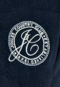 Tissu en polaire marine avec un logo circulaire blanc brodé indiquant "Joules Country Club Established 1989" entourant les lettres stylisées "J C".