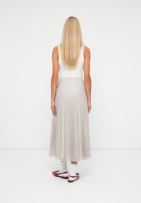 Kvinde med langt blond hår iført en hvid ærmeløs top, sølvfarvet plisseret midi-nederdel, hvide sokker og røde flade sko, stående med ryggen til.