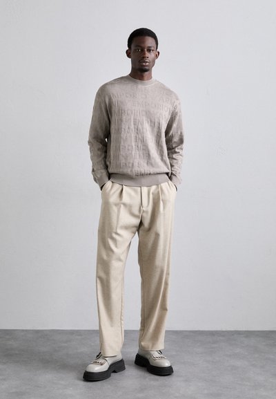 Maglione beige con un sottile motivo a logo goffrato, abbinato a pantaloni beige su misura. Footwear presenta una colorazione grigia con accenti testurizzati e suola spessa.