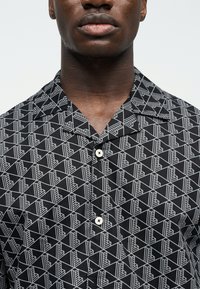 Άνδρας που φοράει μαύρο πουκάμισο με κουμπιά με λευκό γεωμετρικό μοτίβο κύβων, ορατός από τον λαιμό έως το άνω μέρος του θώρακα με φόντο απλό.