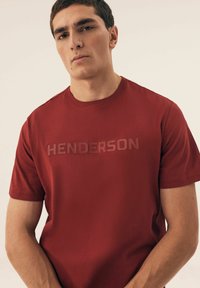 Czerwona koszulka z krótkim rękawem wykonana z teksturowanej tkaniny, z tonalnym logo "HENDERSON" na przodzie. Prosty, okrągły dekolt i luźny krój.