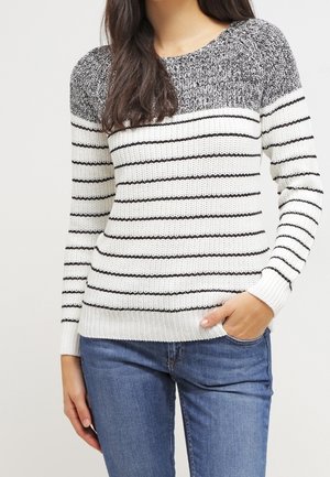 Femme portant un pull en maille avec des épaules gris marbré et un corps blanc avec de fines rayures horizontales noires, associé à un jean bleu.