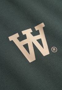 Grüner Stoff mit einem großen, cremefarbenen "W"-Logo und einem eingetragenen Markensymbol, strukturierte Oberfläche mit dezentem Muster.