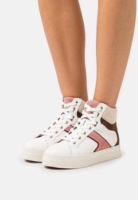 Legs wearing white high-top sneakers with pink and brown suede details and white laces against a plain light background.
