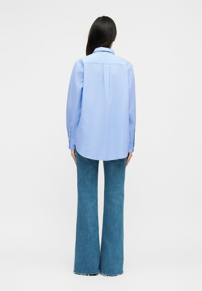 Γυναίκα που στέκεται με την πλάτη προς τα έξω, φοράει ανοιχτό μπλε μακρυμάνικη μπλούζα και μπλε καμπάνα τζιν, μπροστά σε απλό φόντο.