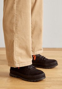 Chaussures noires à surface texturée, avec une semelle épaisse noire et des accents orange. Portées avec un pantalon en velours côtelé beige clair, présentant des rayures verticales.