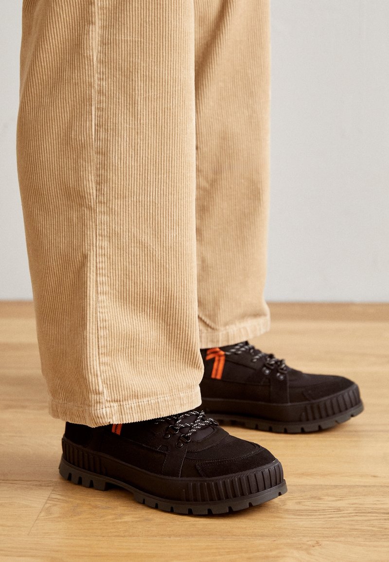 Chaussures noires à surface texturée, avec une semelle épaisse noire et des accents orange. Portées avec un pantalon en velours côtelé beige clair, présentant des rayures verticales.
